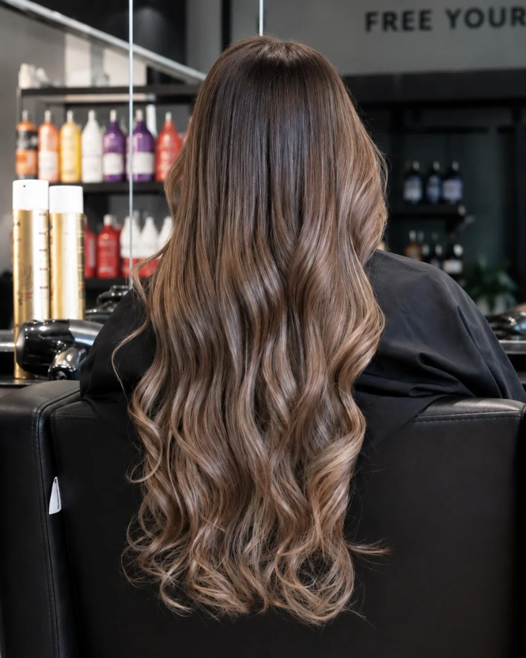 Cheveux longs ondulés aux reflets caramel, vus de dos dans un fauteuil noir